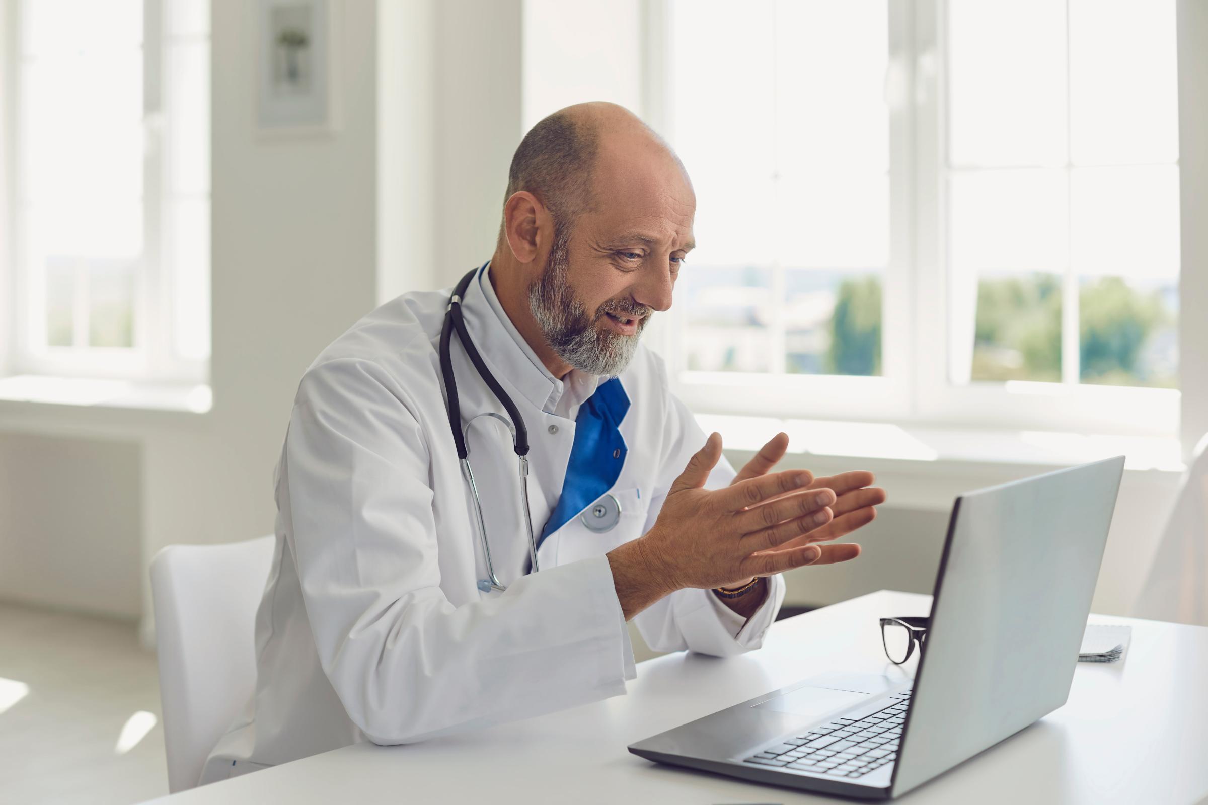 Senior doctor speaks with patient using laptop online video webinar consultation sitting in clinic office.