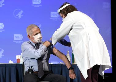 Dr. Fauci is sitting while a healthcare worker administers a vaccine in his upper arm.