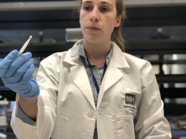 A woman in a white lab coat (Kaitlyn Sadtler, Ph.D., study lead and principal investigator for laboratory testing) uses her gloved hand to hold up a small white microsampling device from the home blood collection kit used in the study.