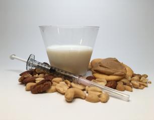 A syringe laying on mixed nuts in front of a glass of milk and wheat crackers with peanut butter spread.