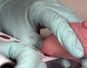 Hand in light blue glove holds newborn’s heel to a white card, which absorbs a spot of blood.