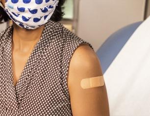 Woman wearing a face mask sitting in a doctors office with a bandage placed over an injection site on her arm.